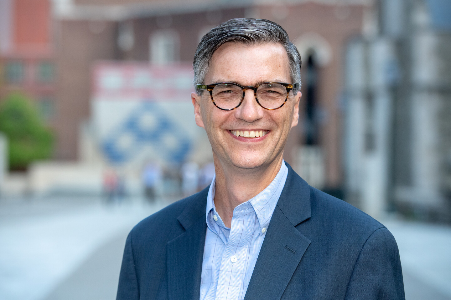 A smiling person wearing glasses in a dark blue shirt and light blue shirt standing outside. 