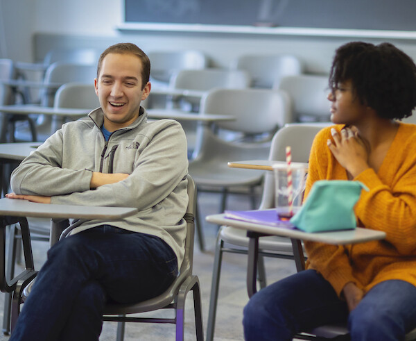 Two people sit at school desks, person on the left is smiling with arms cross as person on the right looks toward him.
