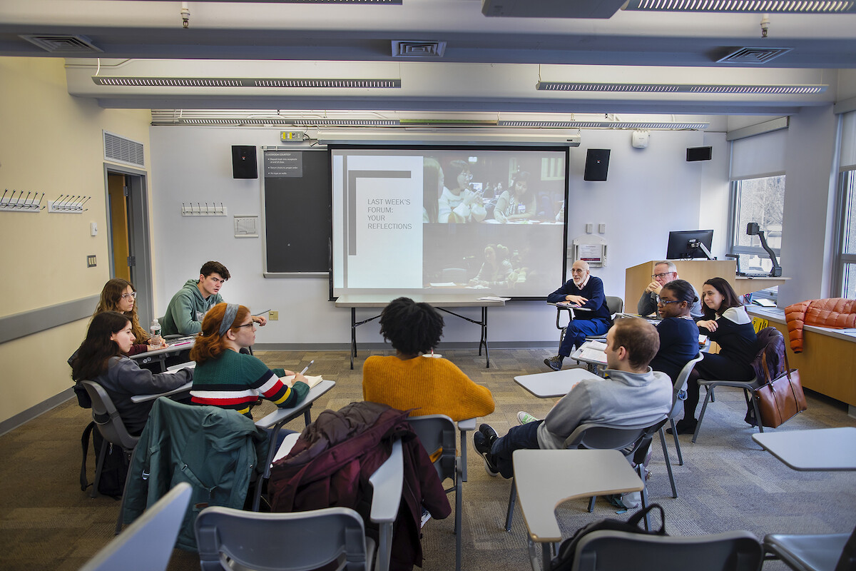 Group of students and teachers sits in desks arranged in semi-circle in front of screen that reads "last week's forum. your reflections."