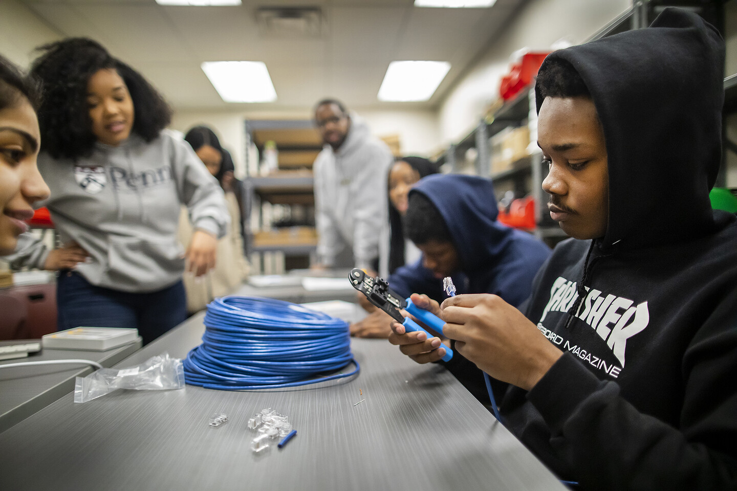 Young man in hoodie uses pliers on coated wire as others look on