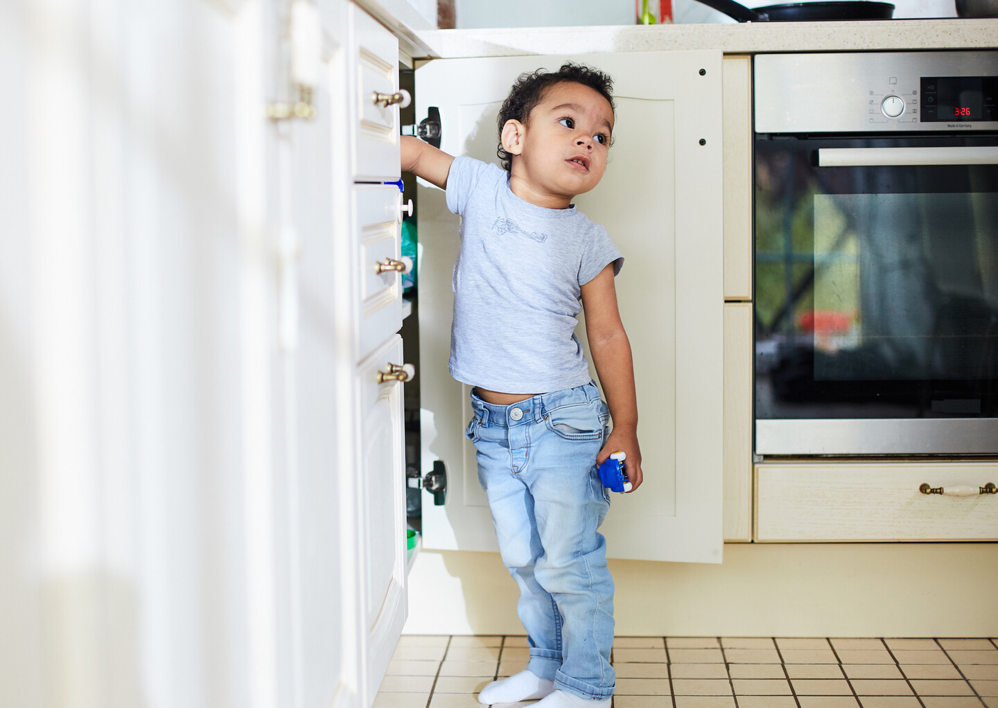 Child standing with a hand in an open cabinet in what appears to be a kitchen. A clock on the stove nearby reads 3:26.