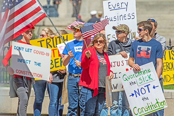 Group of people standing on the steps of a state building holding signs in protest of the state’s stay at home orders due to the pandemic.