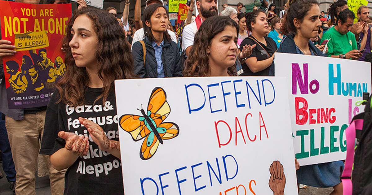 crowd of people at a demonstration holding signs, one reads DEFEND DACA
