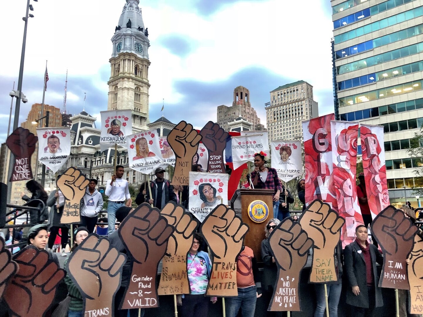 A woman speaks at a podium in front of city hall; all around her people carry signs with black and brown fists and images of slain black trans people. Signs say "trans lives matter" and "say their name."