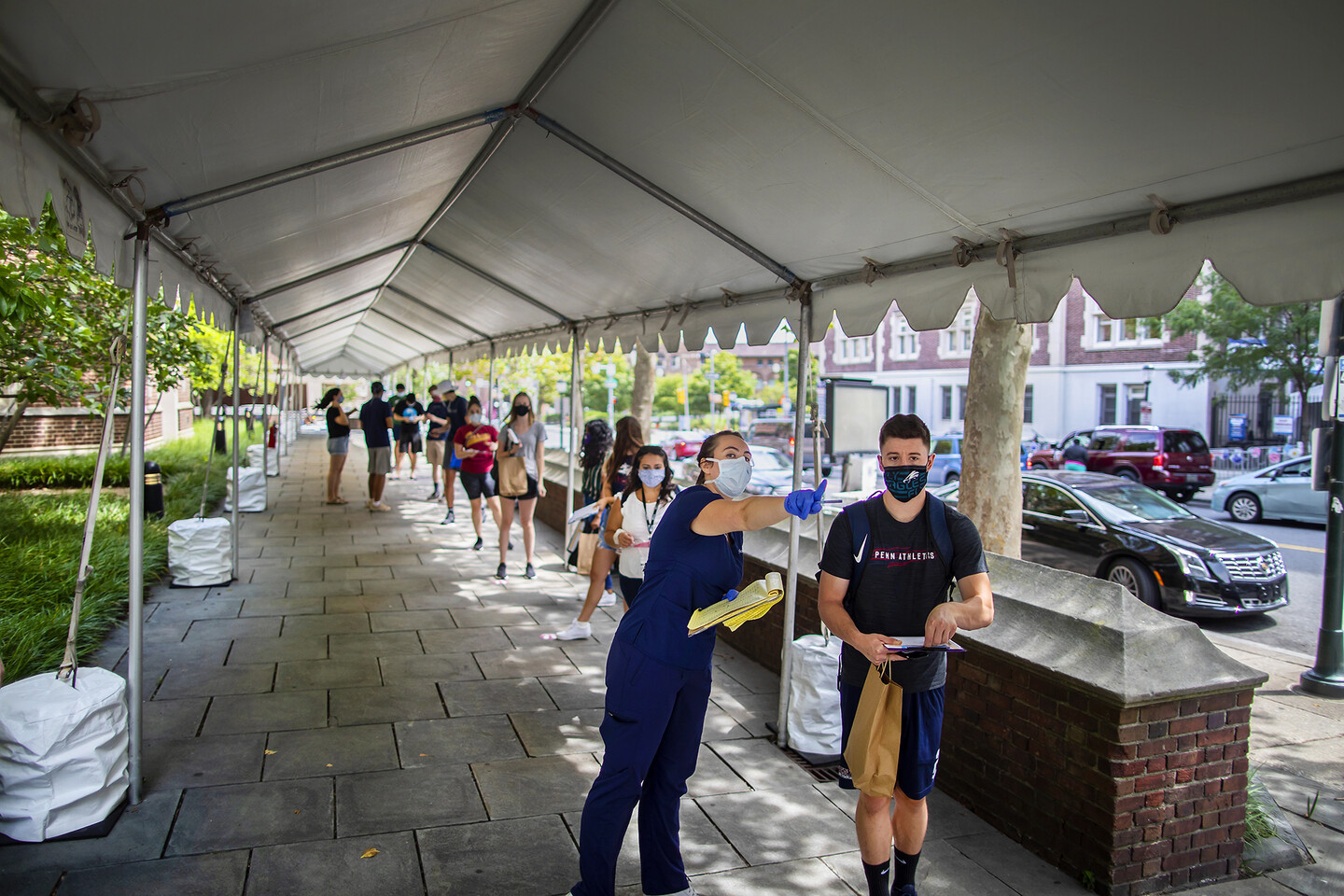 People lined up under a large white party tent, spaced apart, all wearing masks. A person in the front in scrubs, a mask, and rubber gloves points out a location for a person wearing a shirt that reads "Penn Athletics."