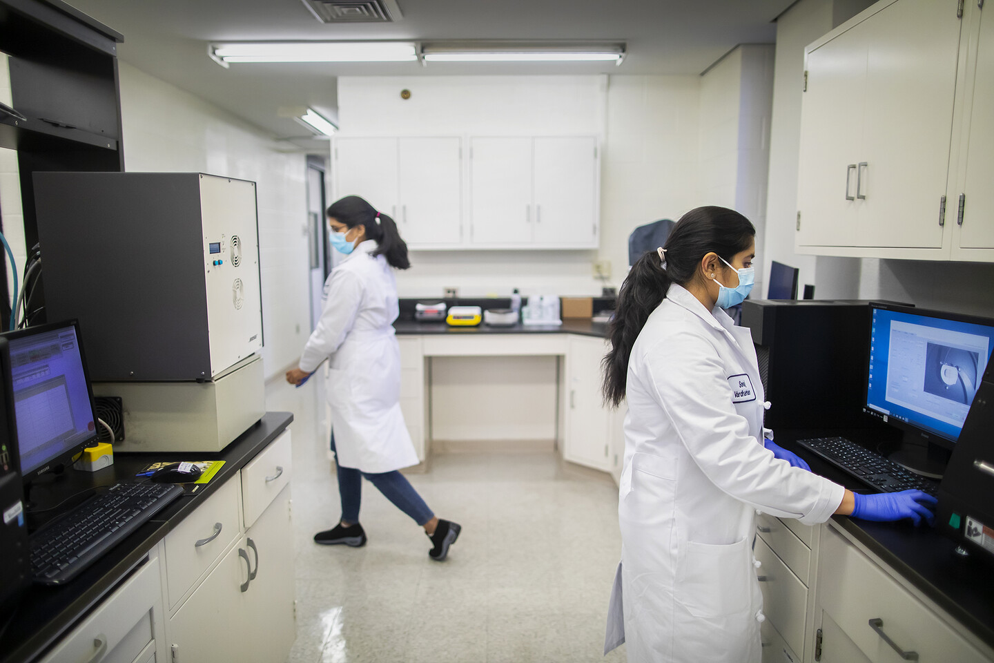 two people wearing masks in a clinical lab, one person at a computer and one walking away