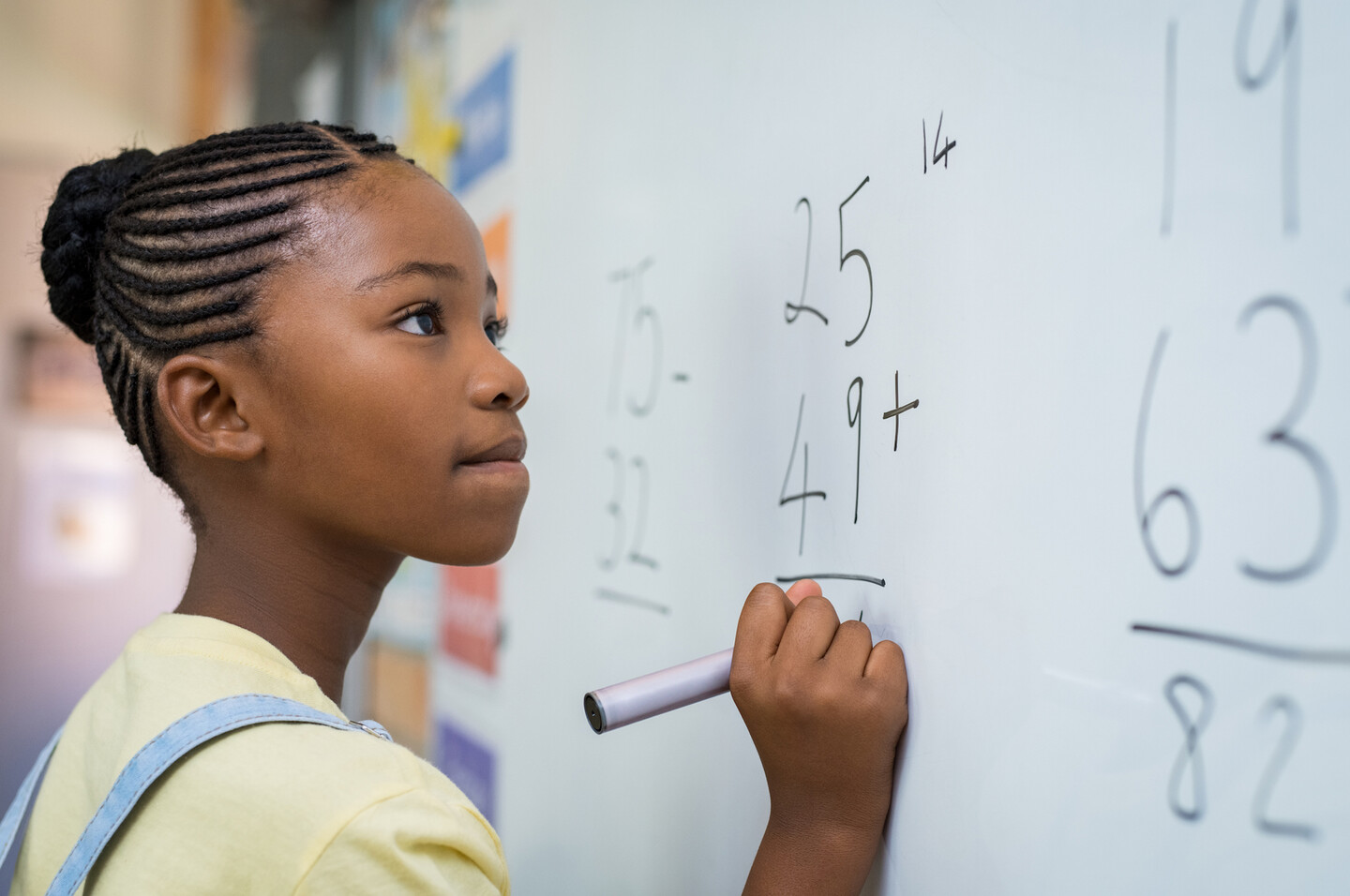 A young girl doing a math problem at a whiteboard. The numbers 25, 49, and 14 are visible.