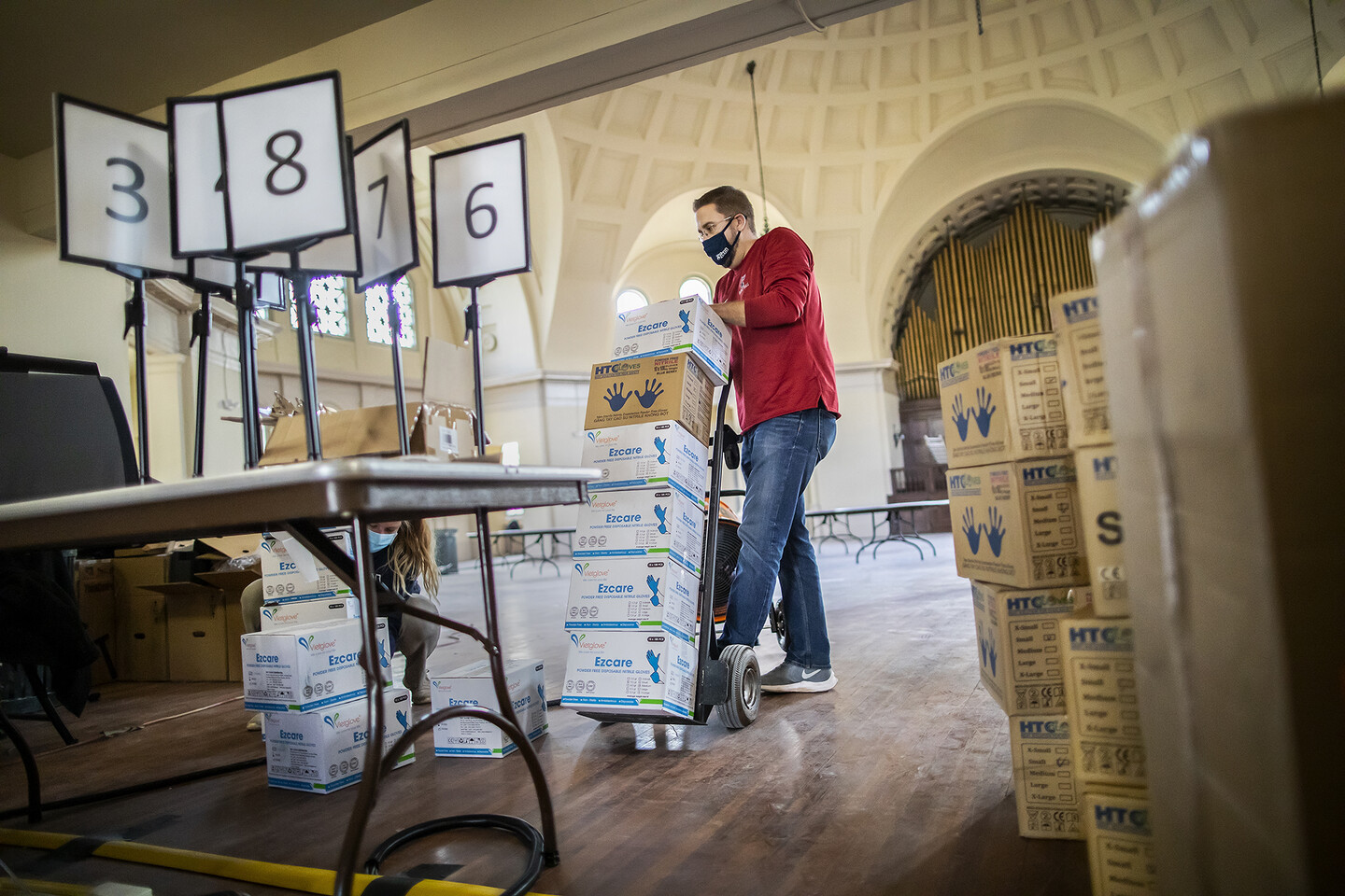 a person wheeling a cart stacked with boxes of gloves and placing them down by other boxes and a table with number placards