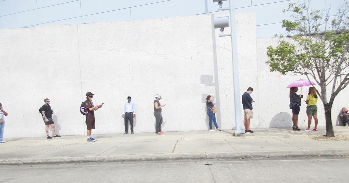 Voters stand in line outside against a white wall, socially distanced and wearing masks.