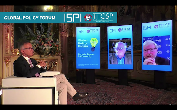 A man in a suit and tie and glasses sits in profile in a white chair as he looks at three video screens in front of him: one showing an illustration of a light buld and the words global policy forum, the middle screen showing a man with glasses in front of a book case and the last screen on the right showing another man with glasses, grey hair and a beard