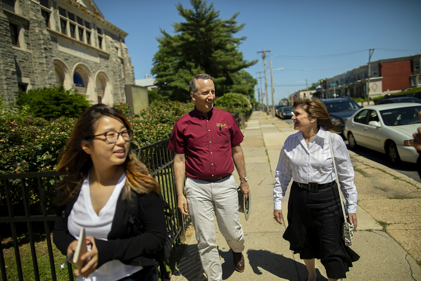 Three people walking along a paved sidewalk, with a concrete building, black iron fence and bushes to their left.