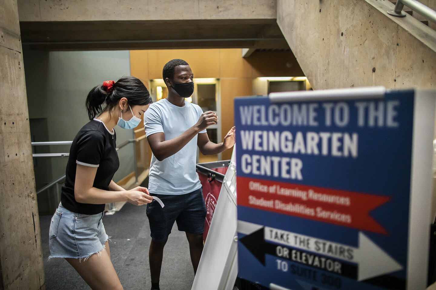 two students in a dorm hallway with a moving cart and a sign