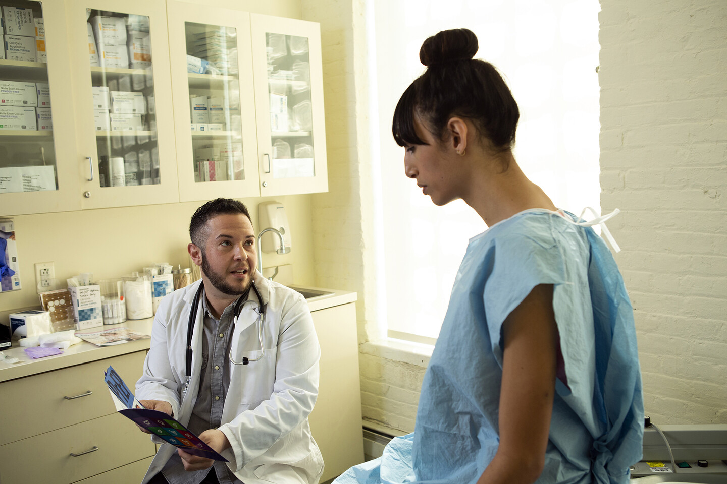 A transgender person wearing a medical gown sits on an exam table speaking to a doctor.