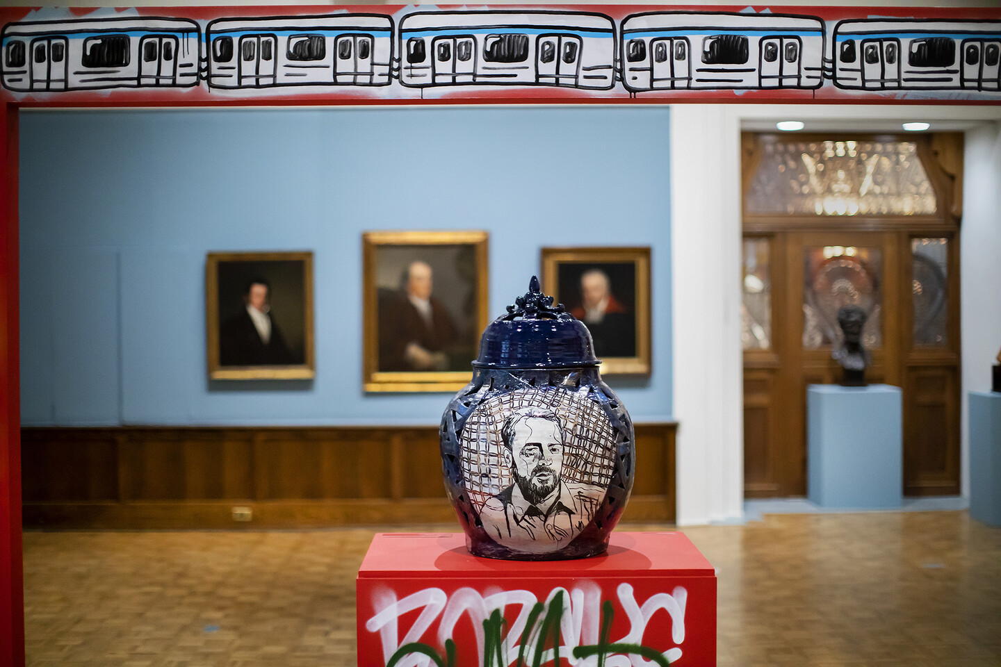 Ceramic pot with a face on it on a pedestal with formal portraits on the wall behind. 