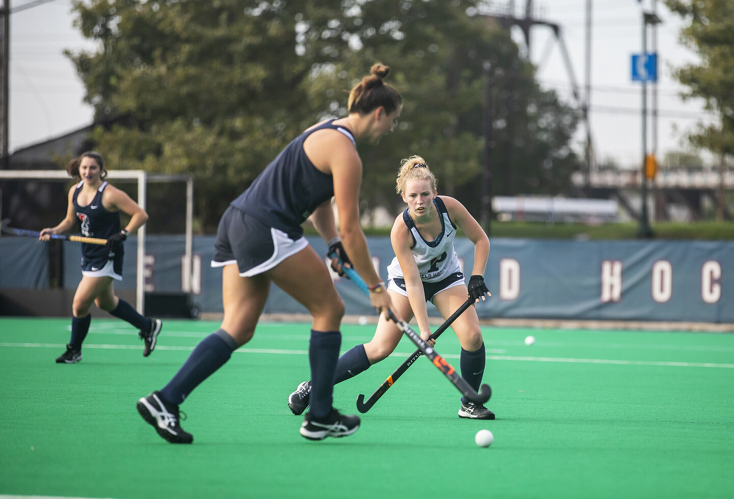 During practice at Vagelos Field, two players, one in a white jersey and one in a blue jersey, battle for the ball with their sticks.