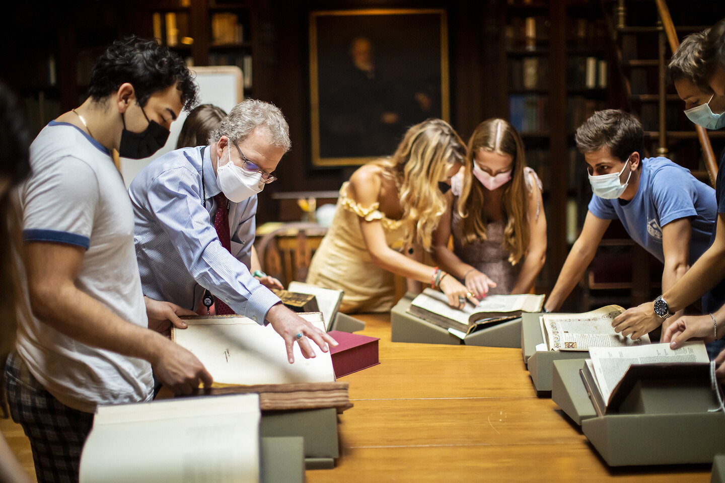 Five people look at different books; a man in a blue shirt leans over to show something to a student