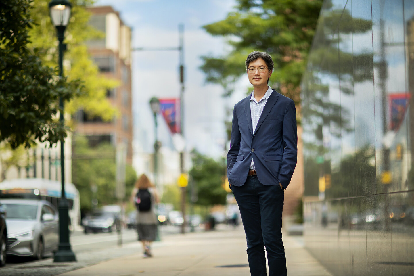 Person in blue slacks and a blue blazer, standing outside on a sidewalk next to a black building, hands in pockets.