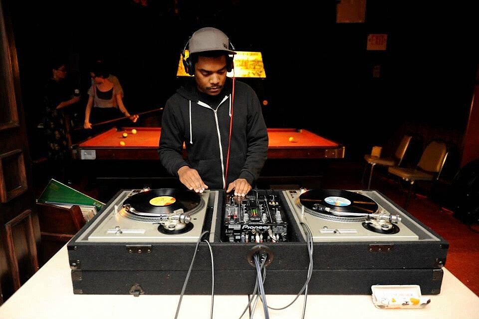 DJ Kid Charlemagne spins records at a turntable, a pool table is behind him.