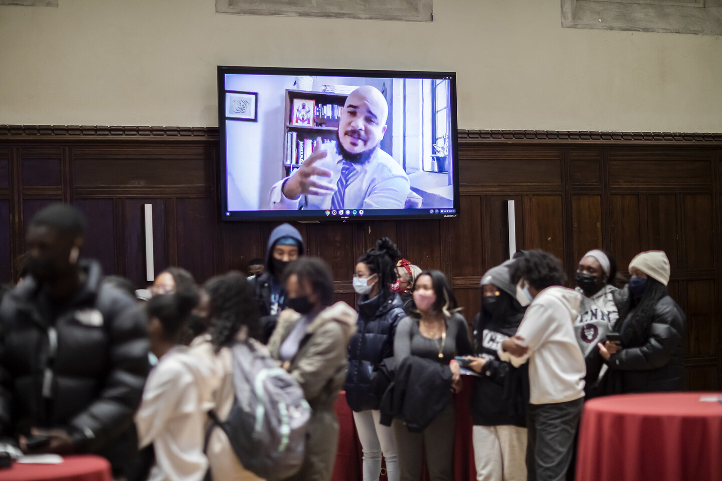 Students mill stand around high-top tables while a man speaks over video