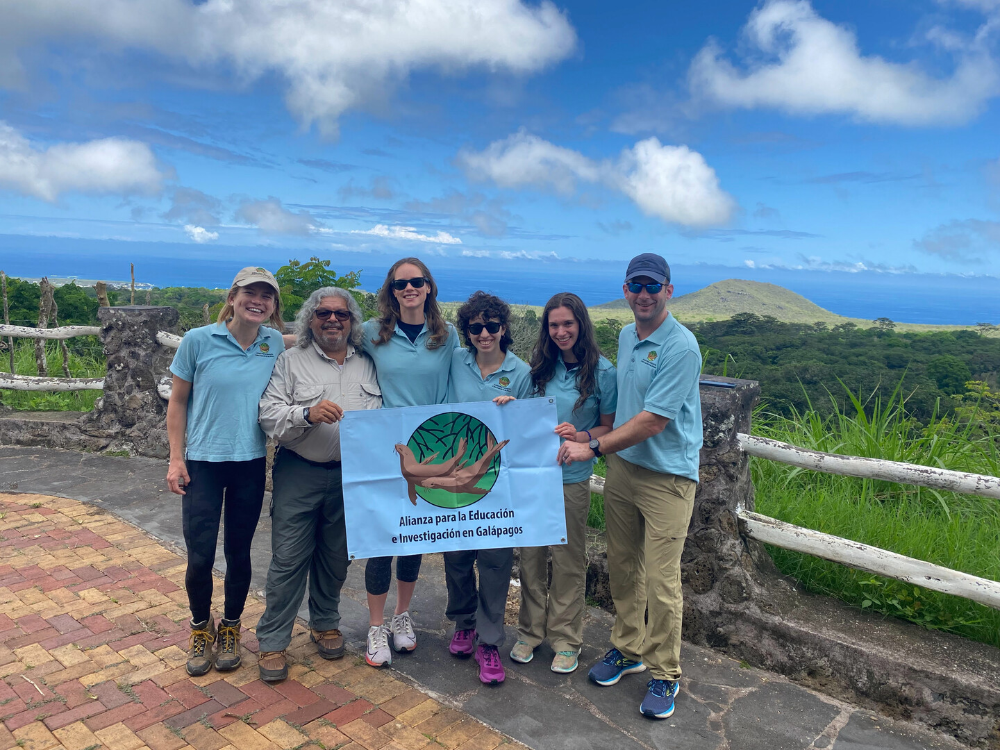 Group poses in a tropical marine landscape holding a sign that reads Allianza para la Educaion e Investigacion en Galapagos