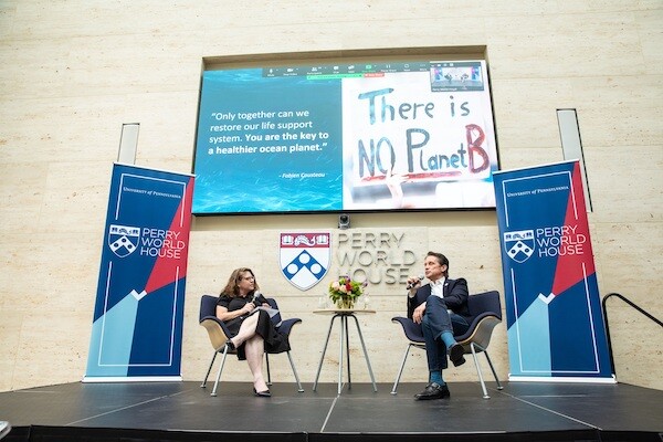 two people sit on the stage at Perry World House with a sign reading "there is not planet b" behind them
