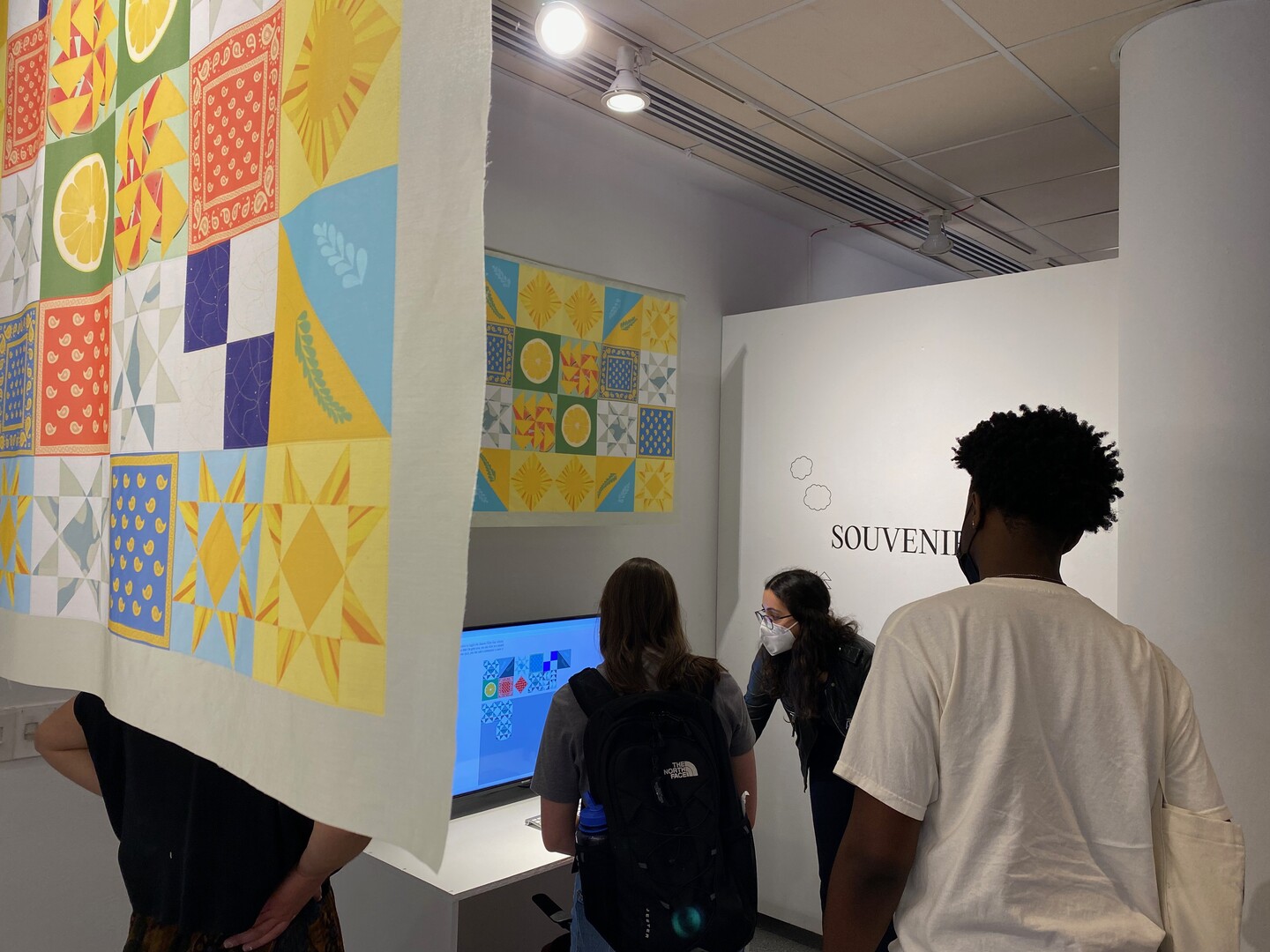 Students standing watching Eleanor Shemtov as she show them her digital quilt project on a computer and her quilt patterns printed on fabric are hanging over the space..