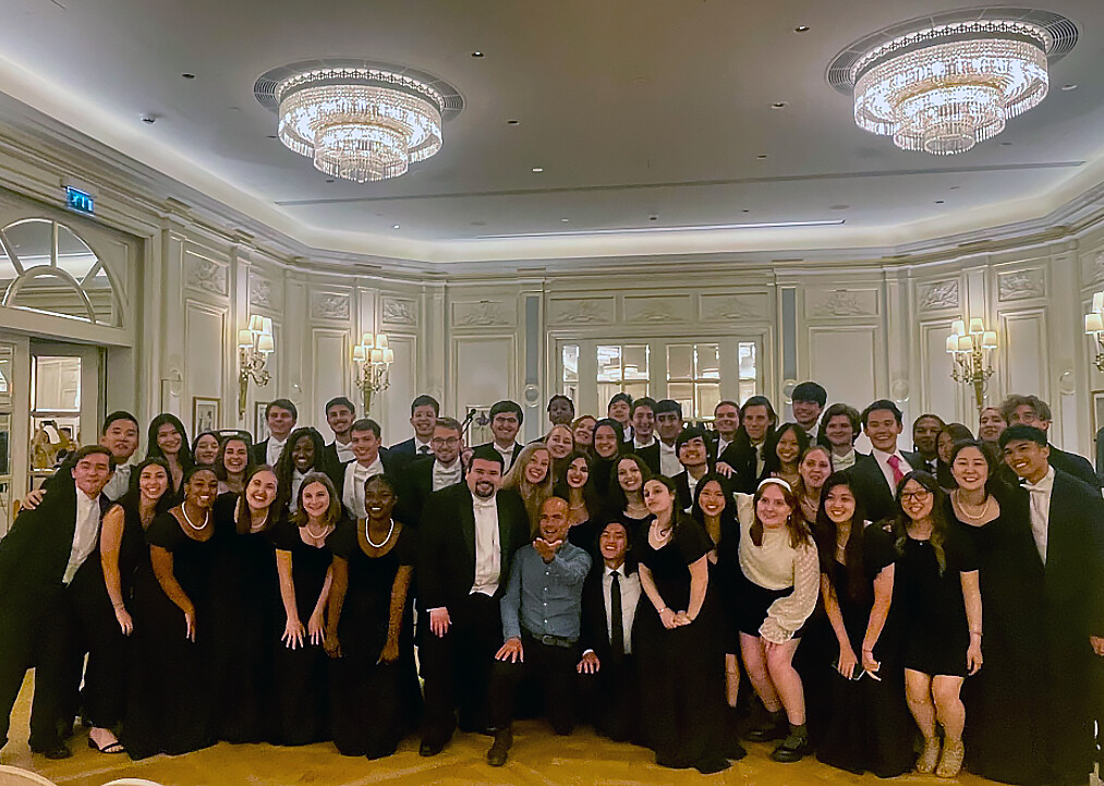 Glee Club members in formalwear gathered together in ballroom under crystal chandeliers