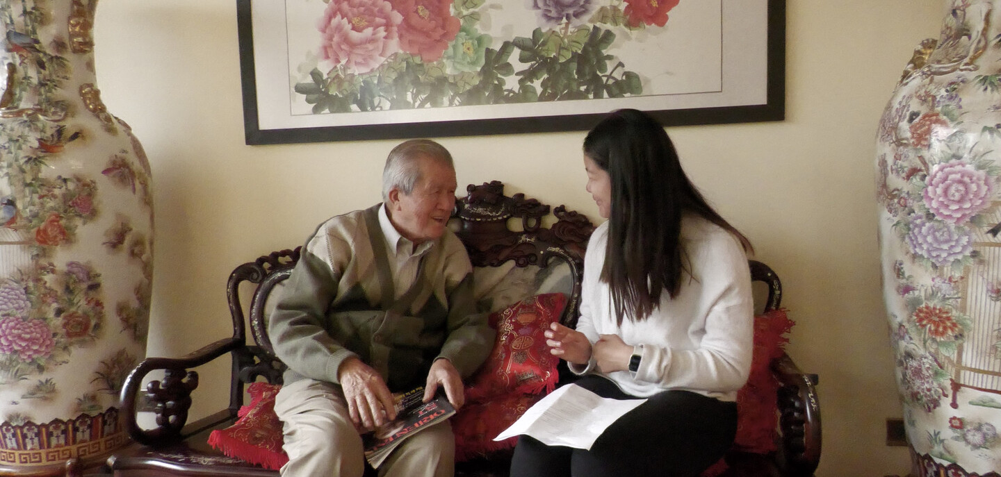 Adrian Ke sits with an elderly man on an ornate bench beneath a painting of peonies.