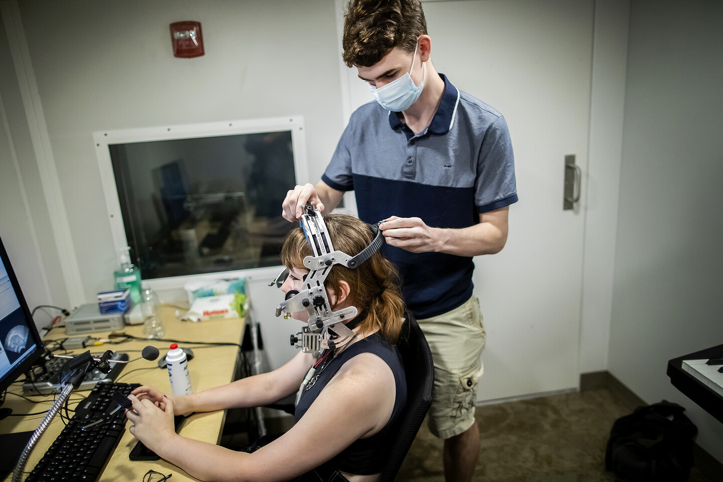 A person standing up adjusting a headset over a person sitting in a soundproof room. Barely visible in front of the sitting person is a computer screen and keyboard. A fire alarm sits above a window behind both people. 