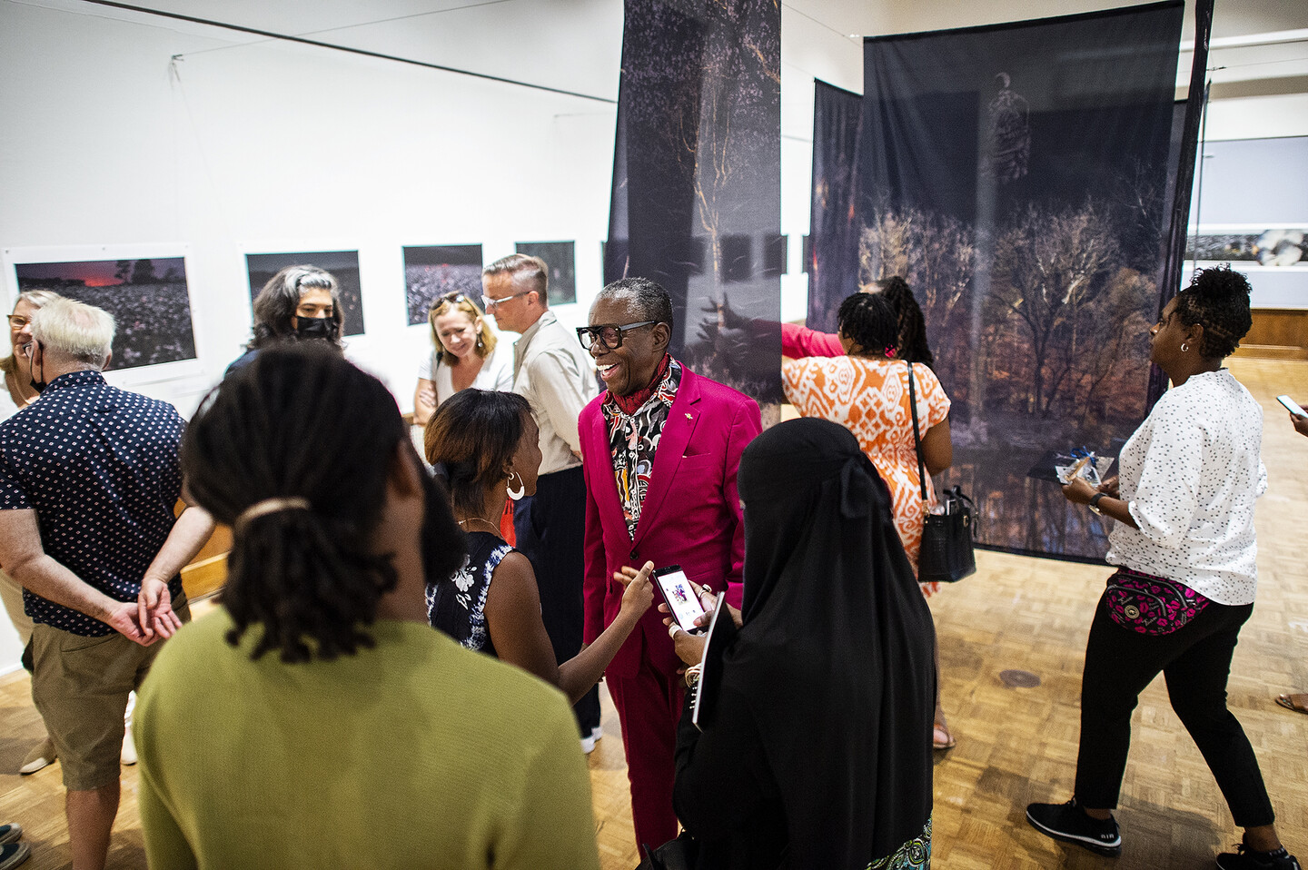 Artist John Dowell stands in the gallery with his artworks behind him surrounded by several people.