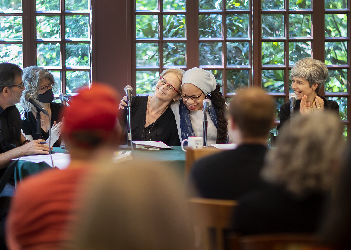 two women hugging while sitting at microphones at a table with people clapping around them