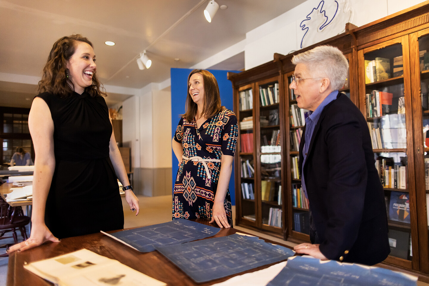 three people talking in archives with bookshelves behind them and blueprints and a journal on a table in front of them 