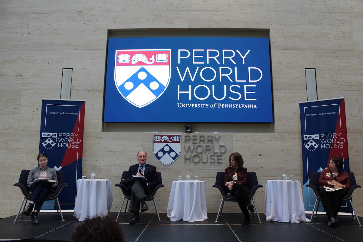 Four people hold microphones, sitting on a stage in chairs separated by round tables with white tablecloths as a sign behind them read Perry World House.