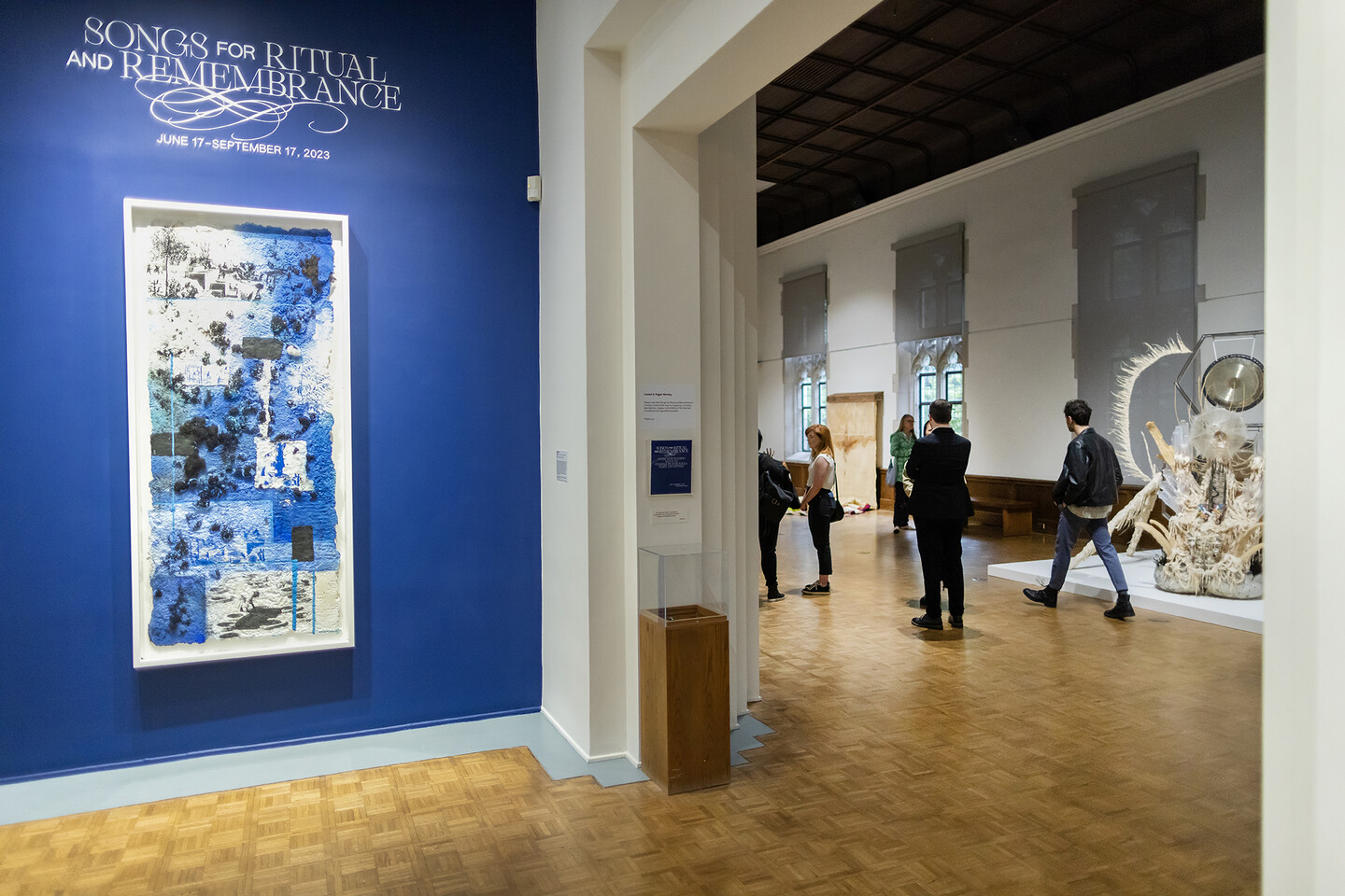 artwork on wall with title Songs for Ritual and Remembrance above and people in gallery behind 