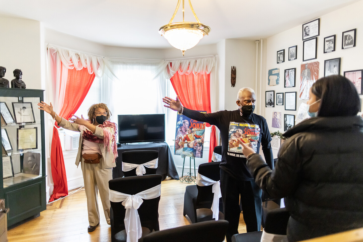 A room with black chairs in front of a television, surrounded by movie posters, albums, and framed photographs. Two people gesture to something on the wall. 