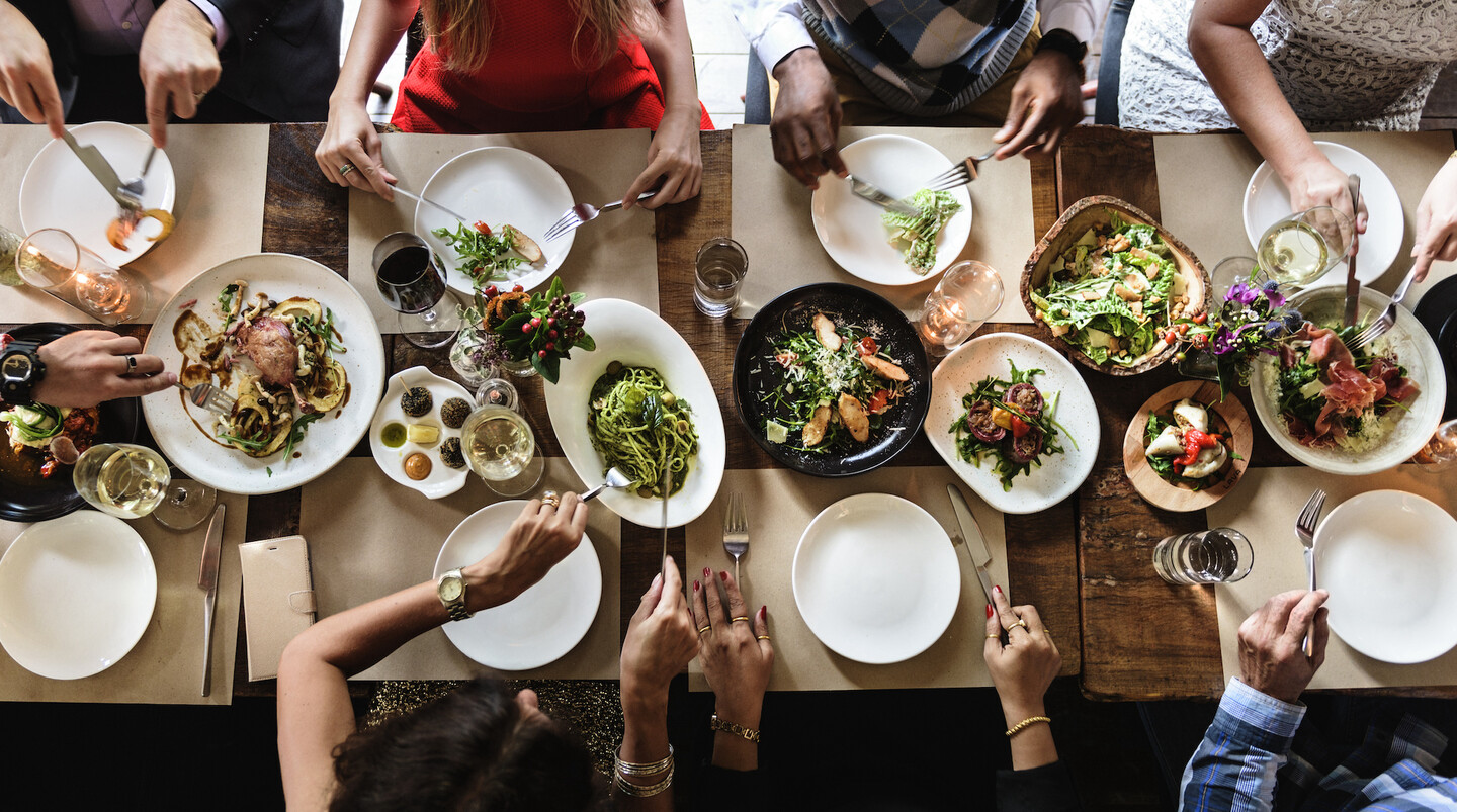 Overhead image of a table set with food and diners hands and arms cutting food and and eating.