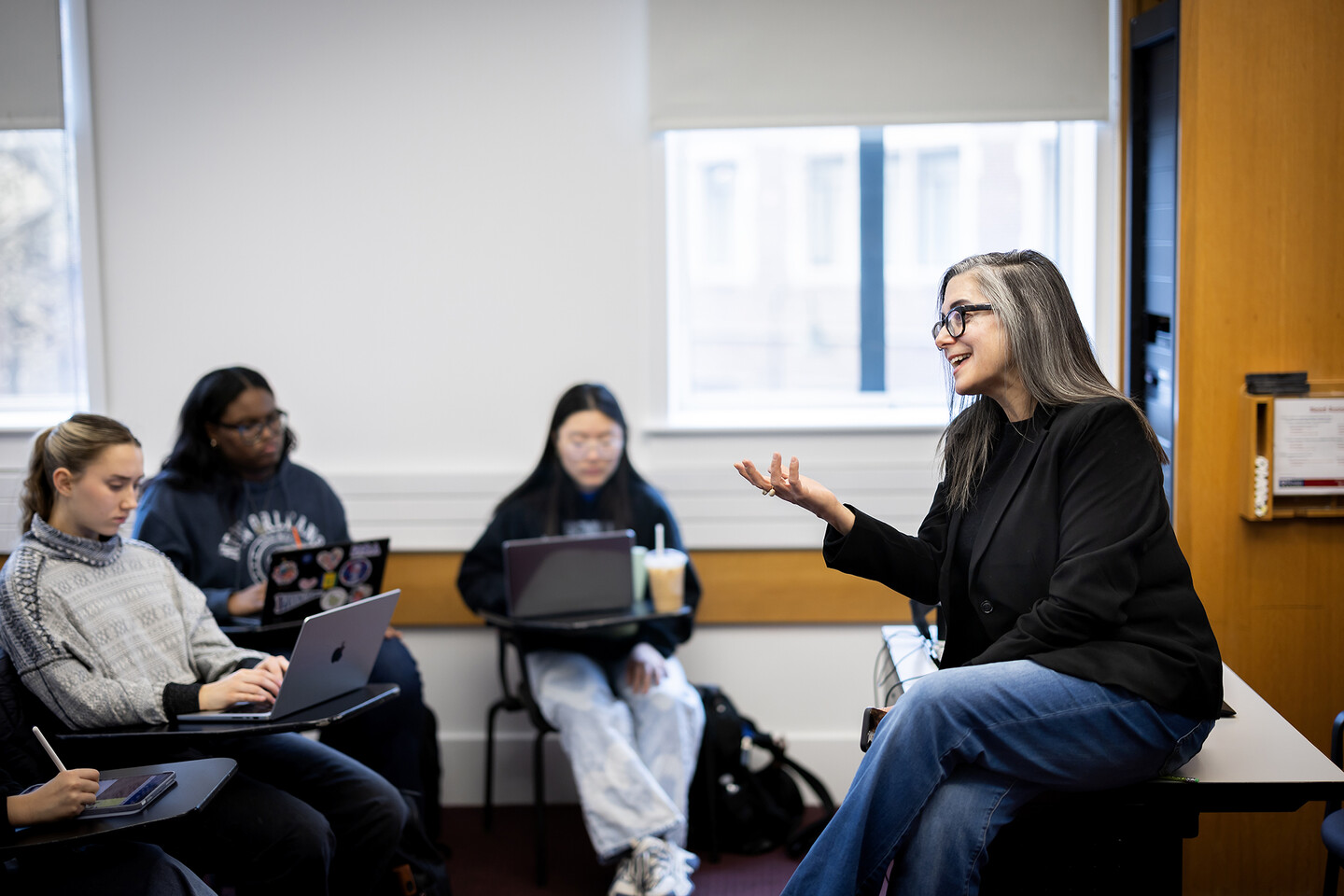 Melissa Jensen sitting on a desk gesturing with her hand to the students in the front row of the class. 