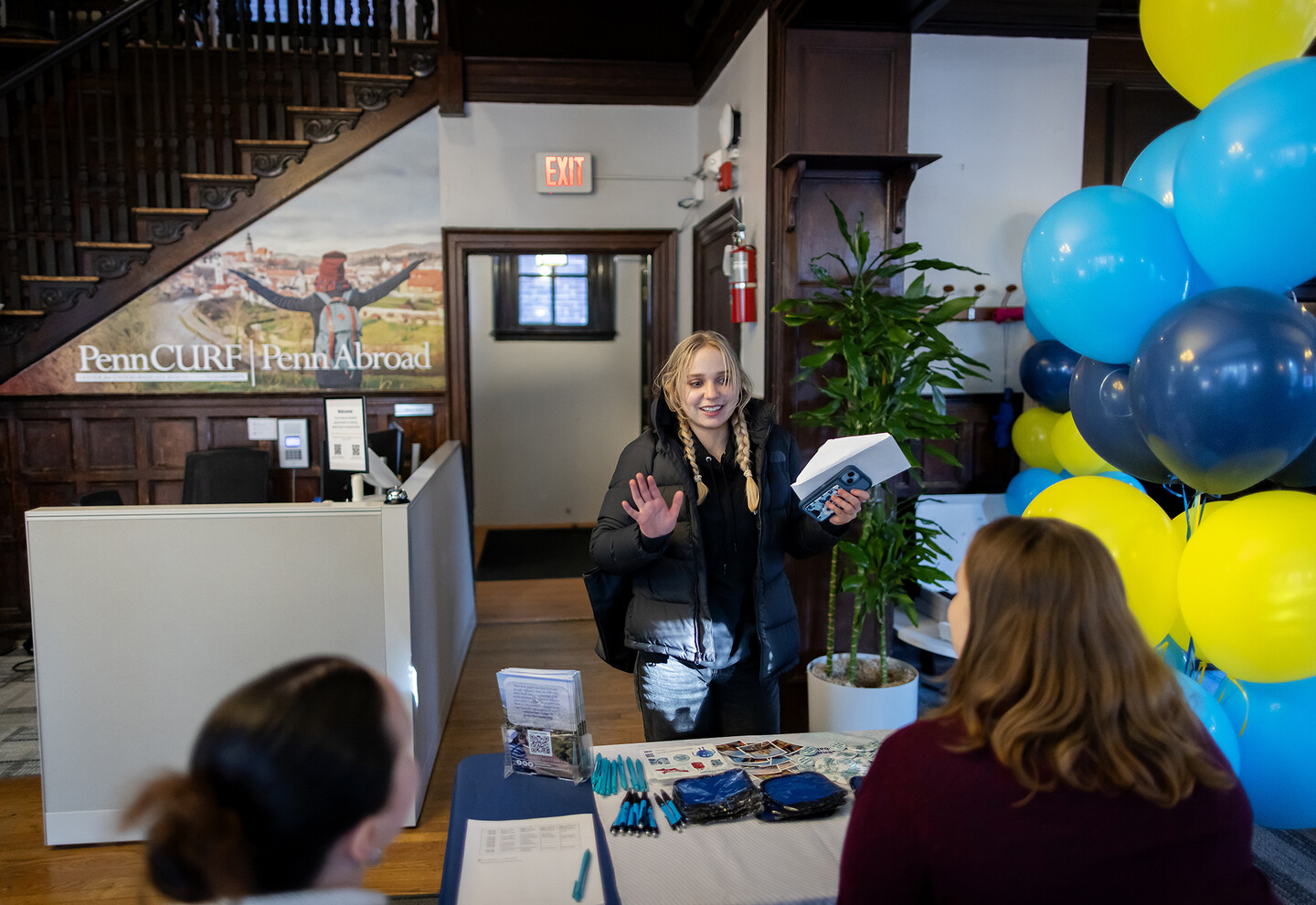 A girl with two braids waves to two people seated at a table with blue and yellow balloons.