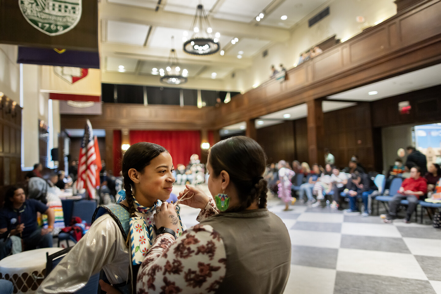 Two women, one tying a bow on the other's regalia