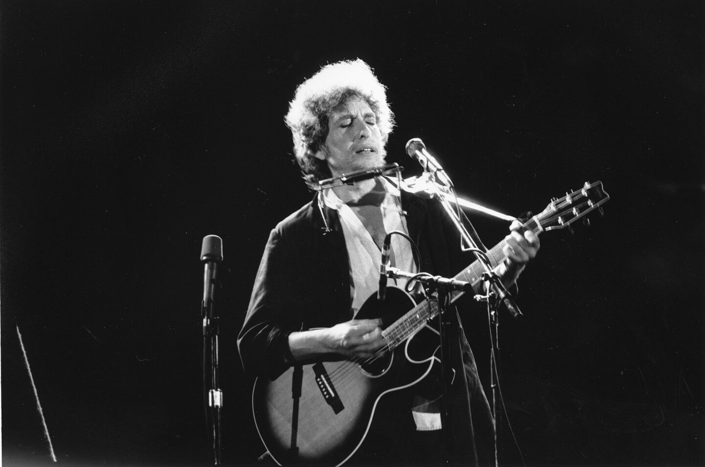 Bob Dylan on stage playing his guitar with his harmonica on a holder near his face.