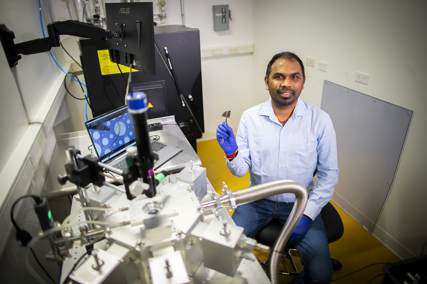 A seated researcher holds a metal pinching tool clasping a metallic device.  