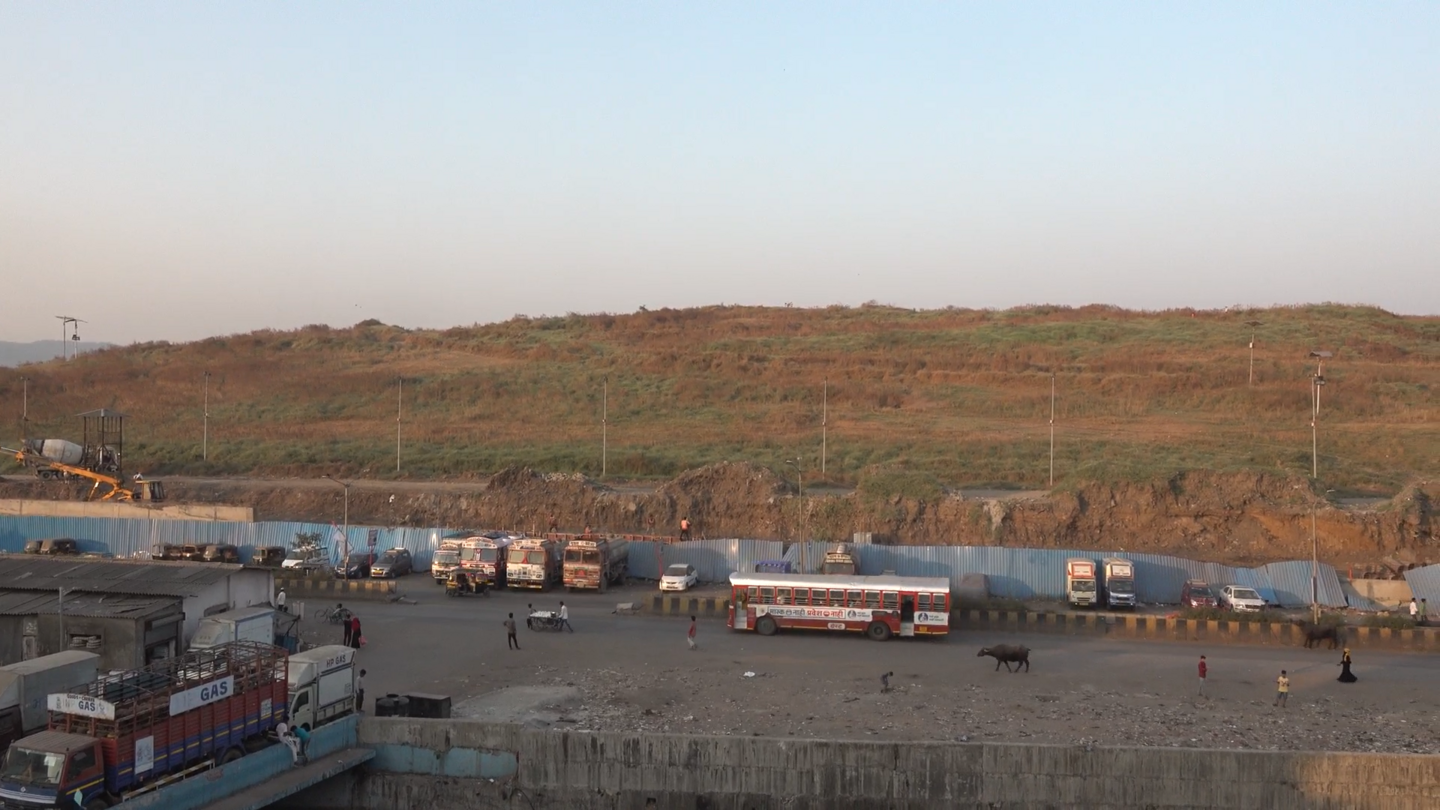 In the foreground, buses, cows, and people idle along a dusty road. Fenced off in the background are the brownish green hills of the unused dumping ground. 
