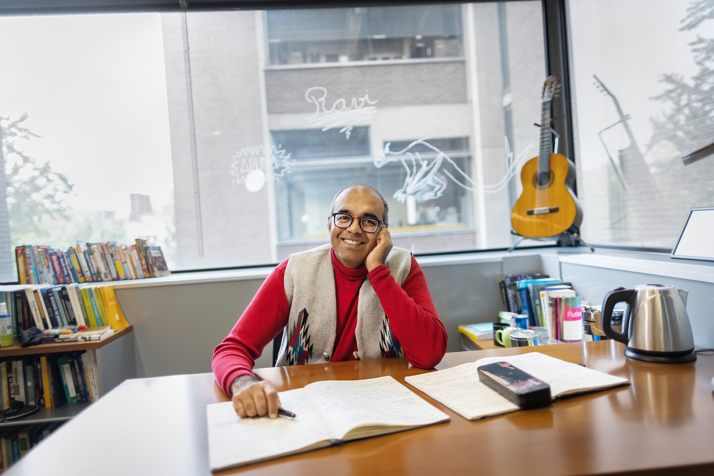 Vijay Balasubramanian sits at his desk with a notebook spread out in front of him. The office has books, an electric teakettle, and a guitar in the window. 