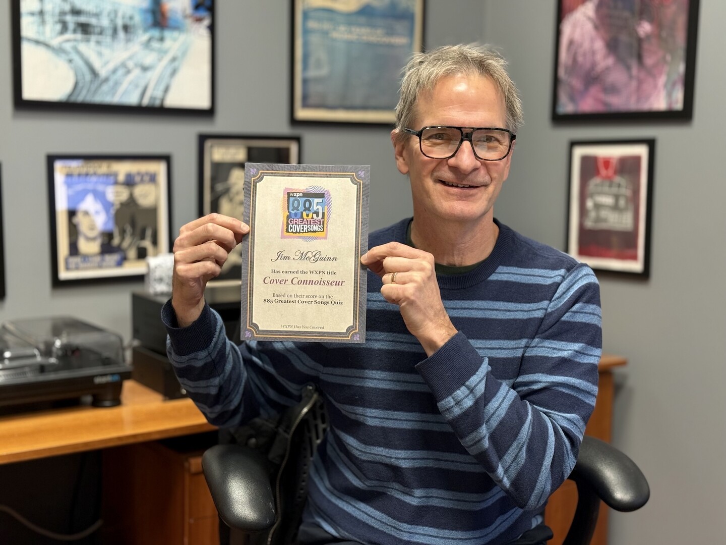  Person seated in an office holding a certificate that reads “Cover Connoisseur” with WXPN branding, surrounded by framed posters on the wall.