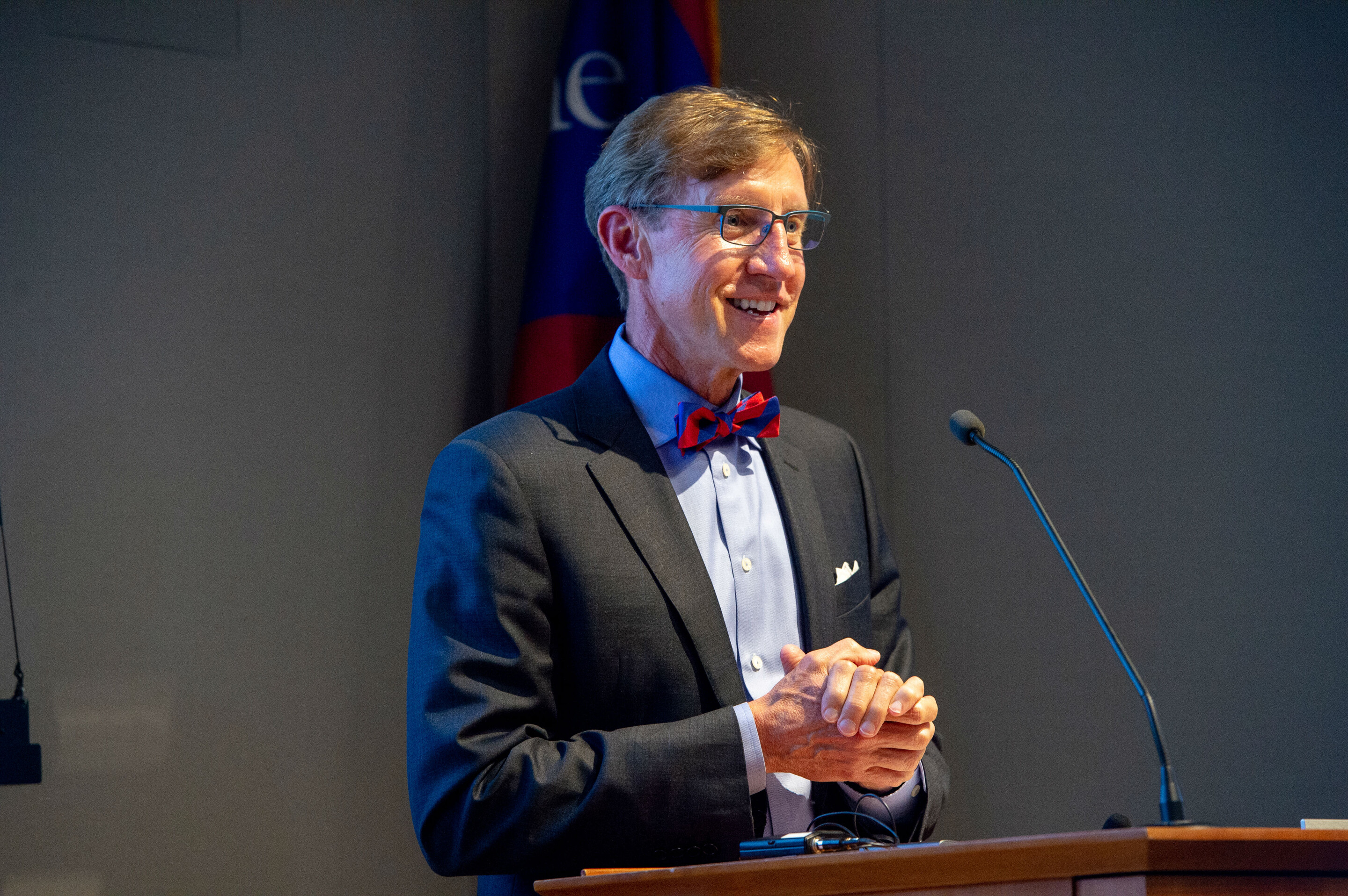 Man at a podium in a grey jacket, blue shirt, and red-and-blue bowtie. 