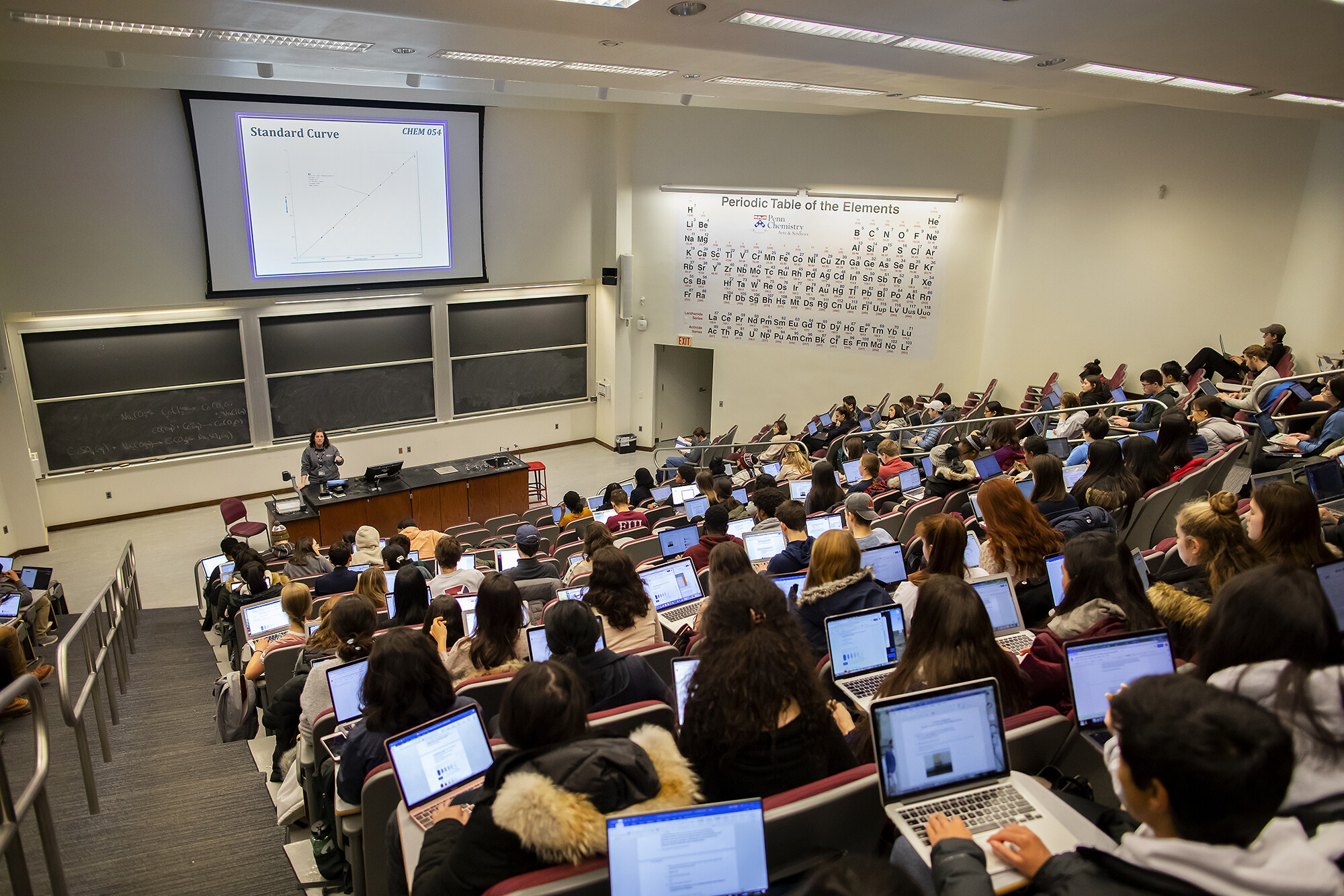 students in a classroom working on computers with a teacher at the front of the room by a chalkboard, at the side is a large periodic table