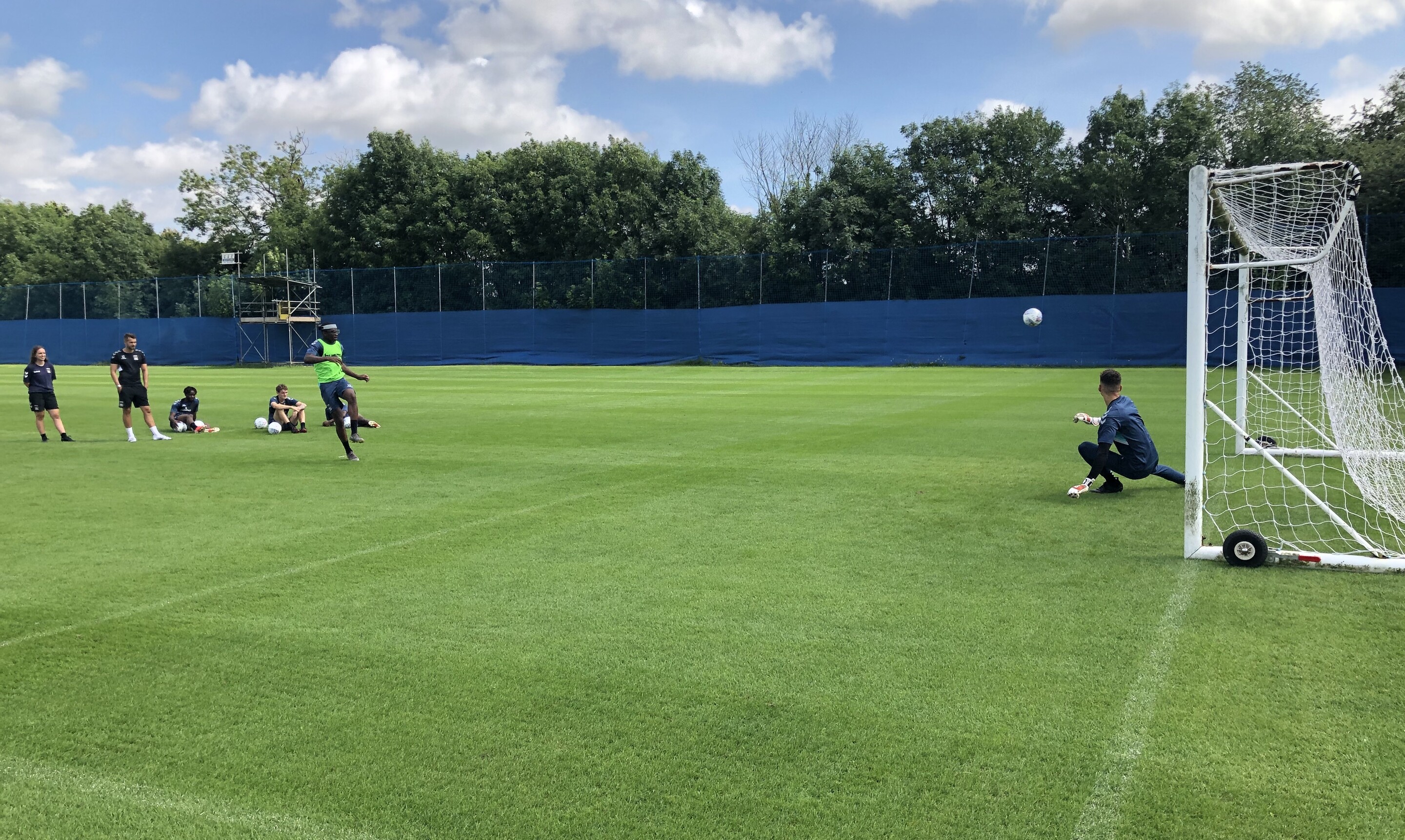 Soccer players on a field on a beautiful day. Four players sit as a fifth, wearing a green vest, kicks the ball toward a goal and a goalie. The ball is in mid-air. 