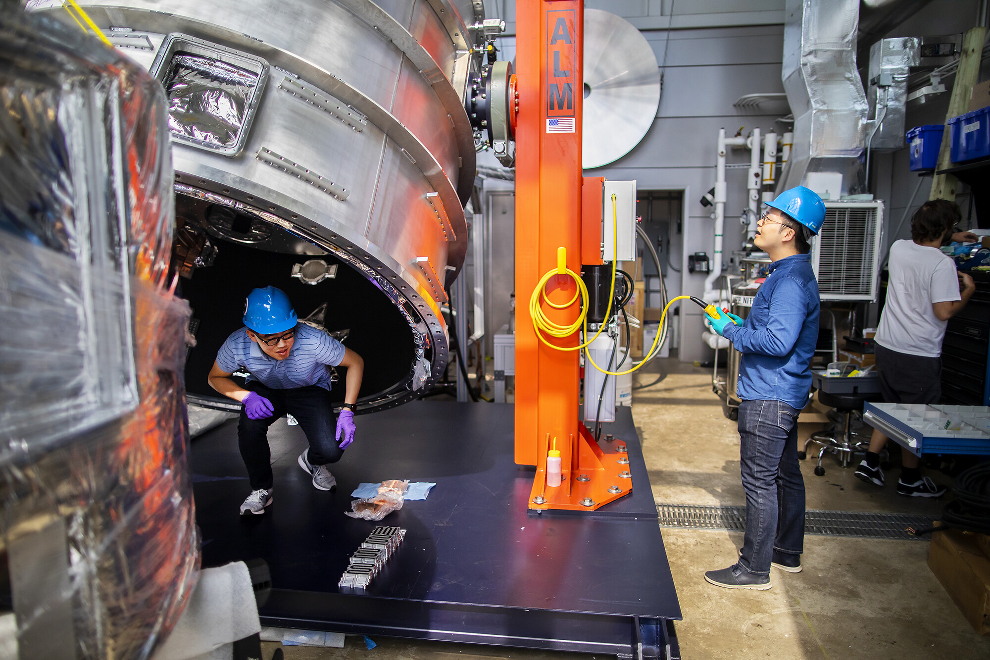 two people in hard hats, one crouching as he steps outside of a large metal shell, the other holding a control device while looking up at the structure