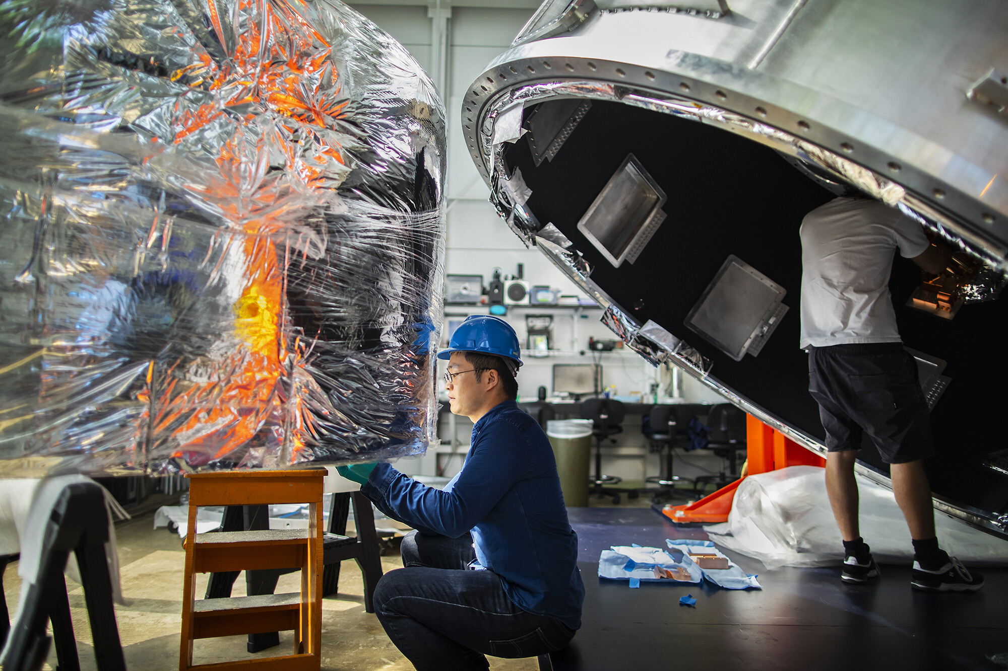 a person in a hard hat sitting on the floor working with a ring of insulation behind a massive metal cyllinder, which a person is standing inside and working on in the background