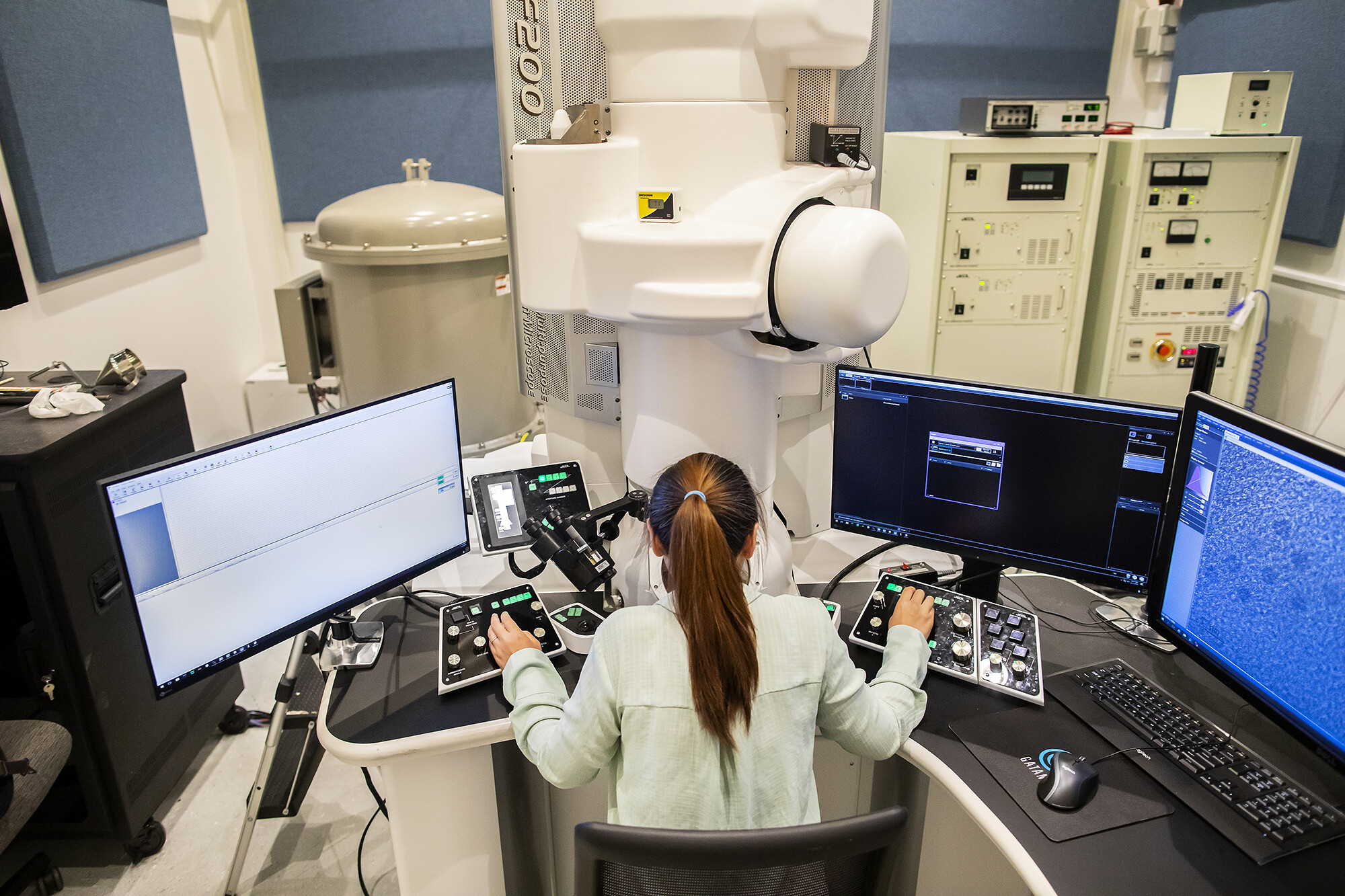 lee adjusts knobs that connect to a massive white microscope, surrounded by multiple computer displays